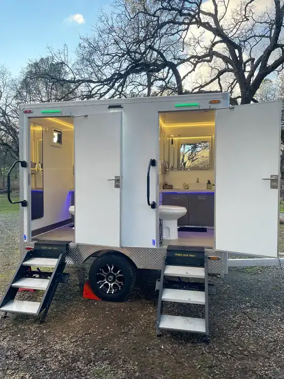 Two-stall restroom trailer lit up at dusk with steps down to the lawn